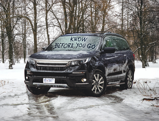 A black SUV covered in snow with 'Know Before You Go' written on the windshield, emphasizing the need to stay alert as winter conditions can change rapidly.